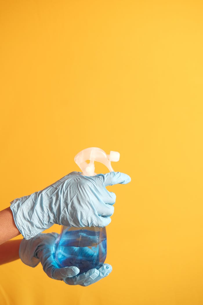 Hand in blue glove holding a cleaning spray bottle against a yellow background.