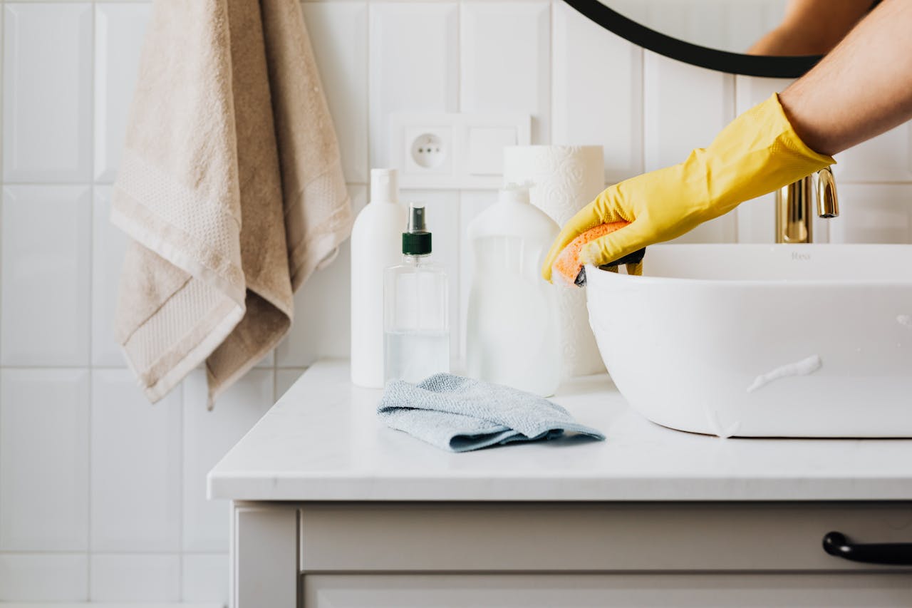 who-we-are Person cleaning a bathroom sink with yellow gloves and cleaning products on a countertop.