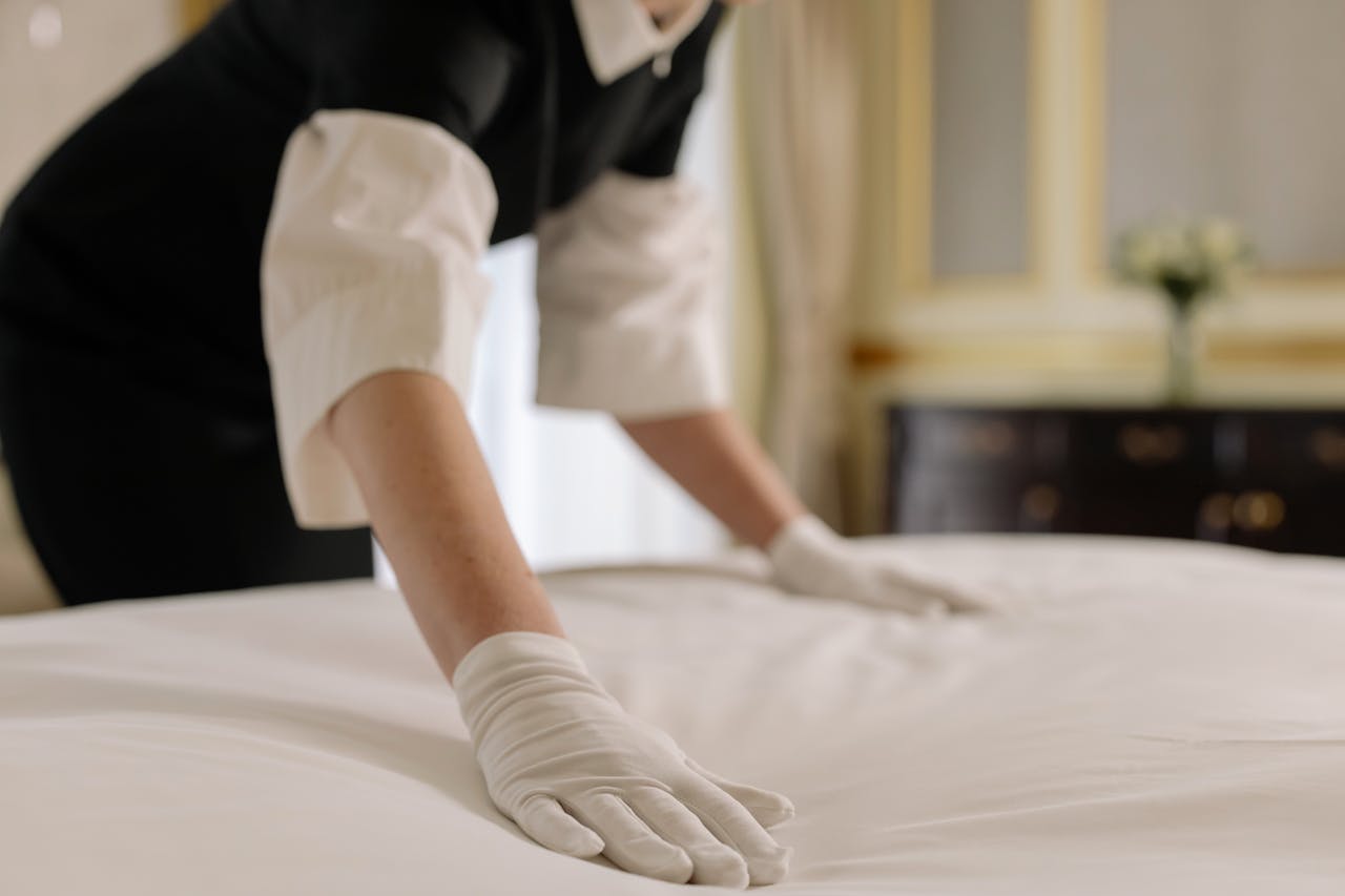 about-us A housekeeper straightening the bed linens in a well-appointed hotel room.